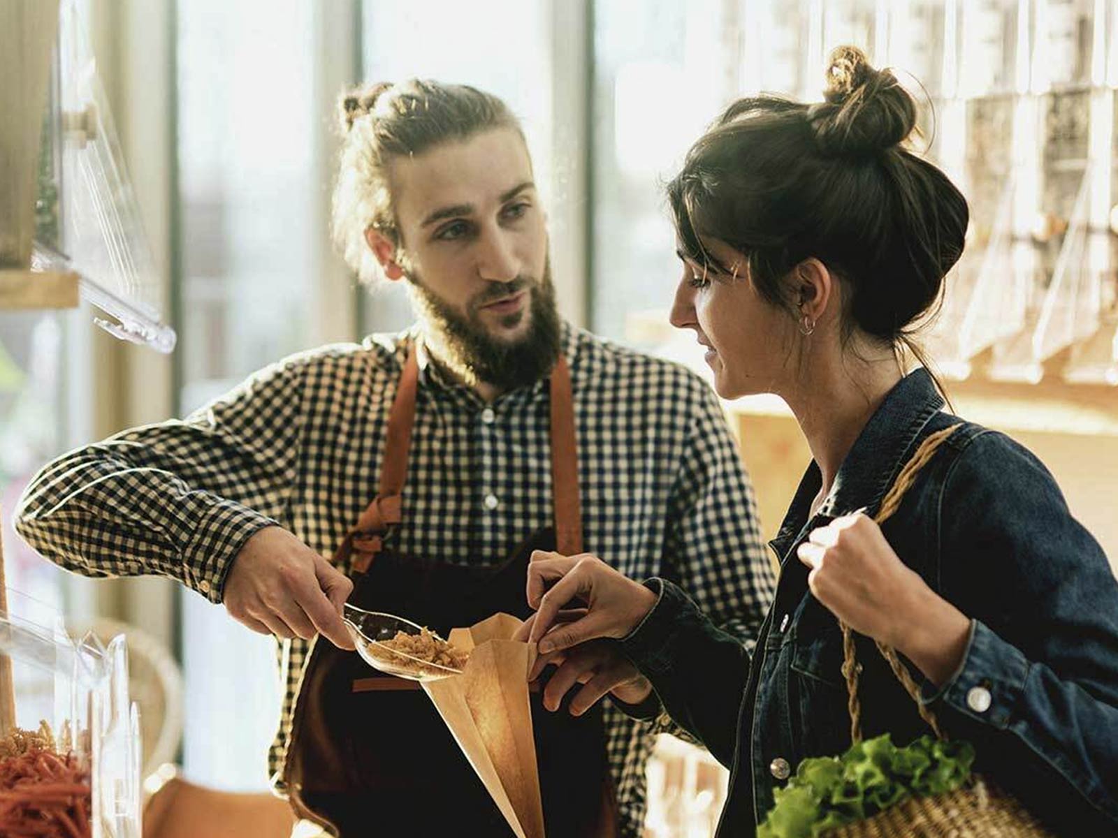 Shop worker in a checkered shirt and apron handing a wrapped food item to a customer at a market counter.