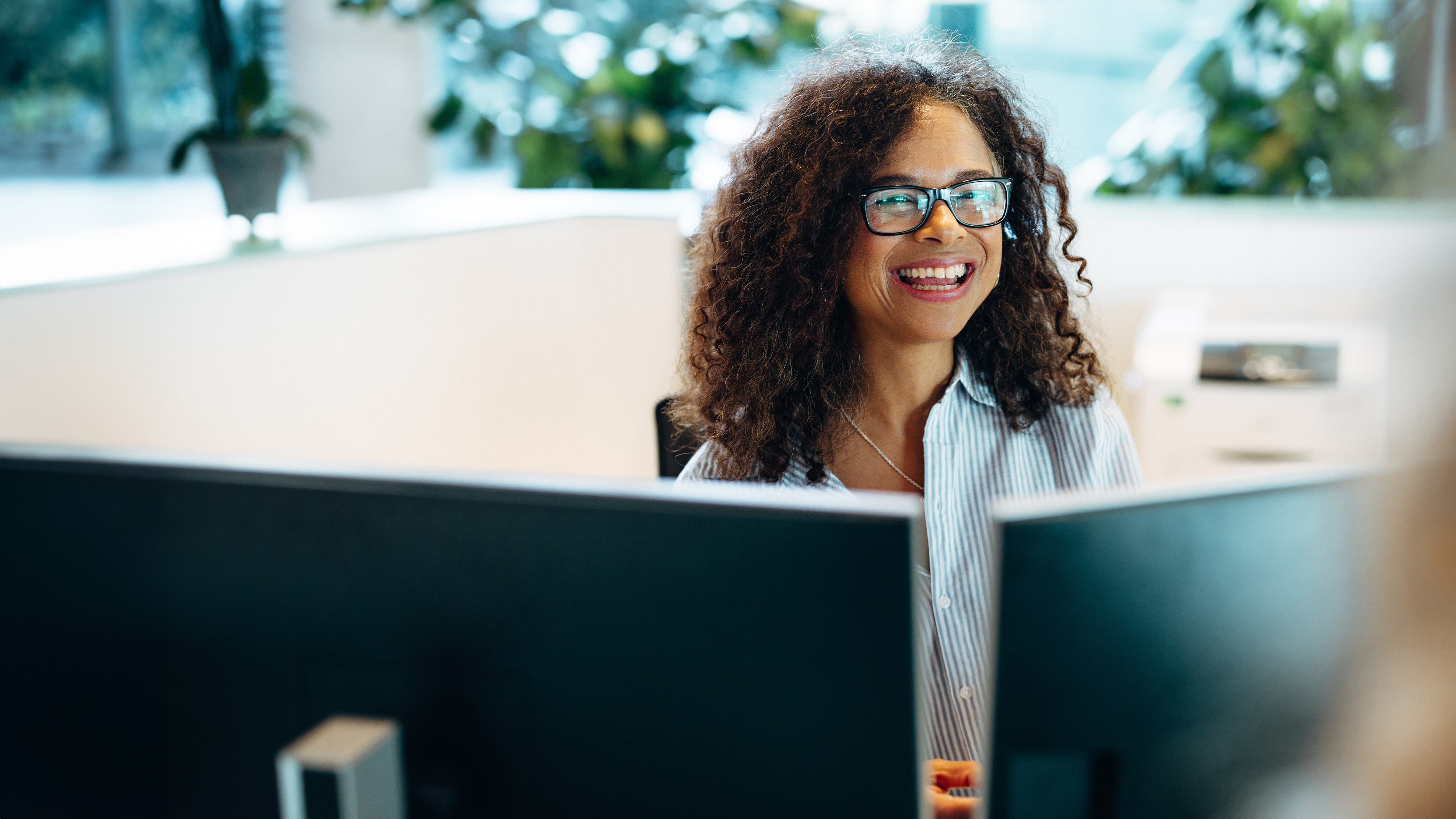 Woman with curly hair and glasses smiling while working at her desk in an office cubicle