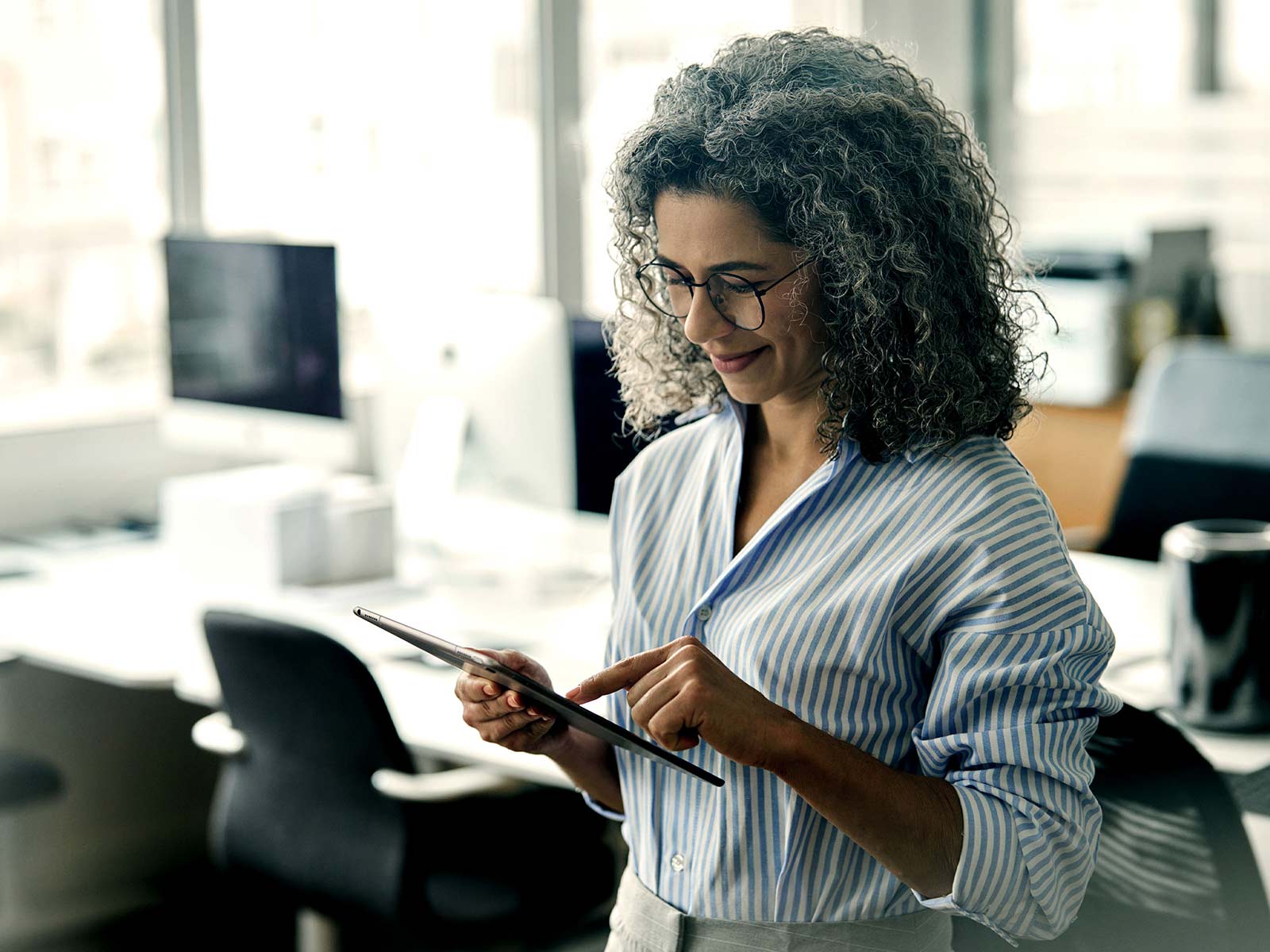 Woman with curly gray hair using a tablet while standing in a modern office
