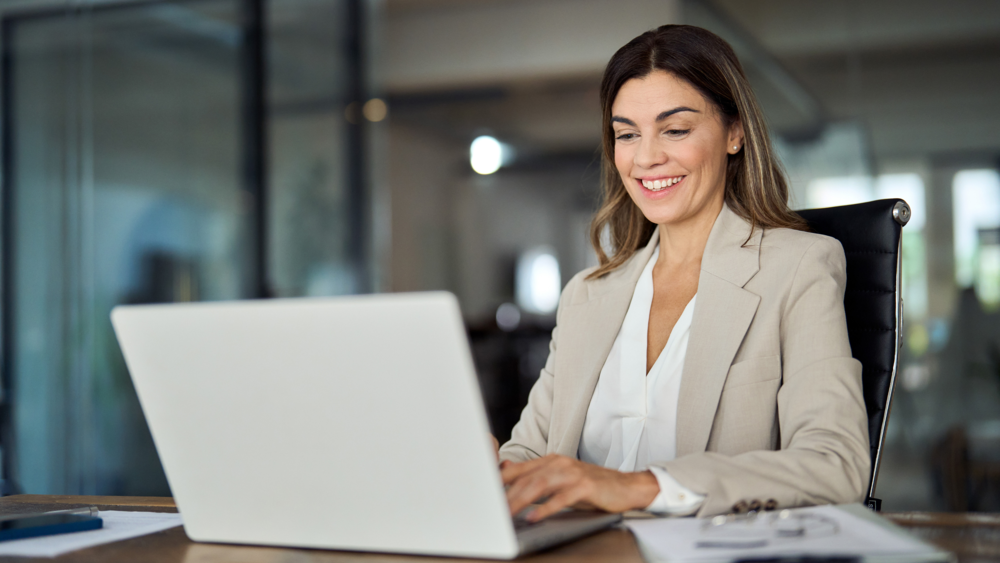 Smiling businesswoman in a blazer working on a laptop at an office desk
