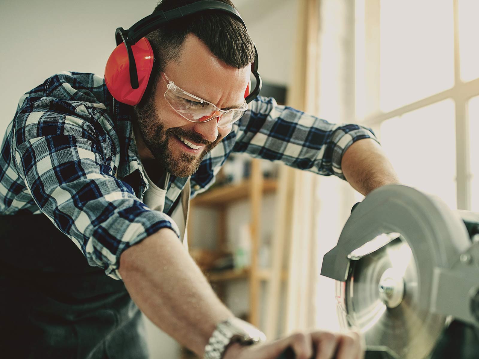 Man in a plaid shirt, safety glasses, and ear protection smiling while operating a circular saw in a workshop.