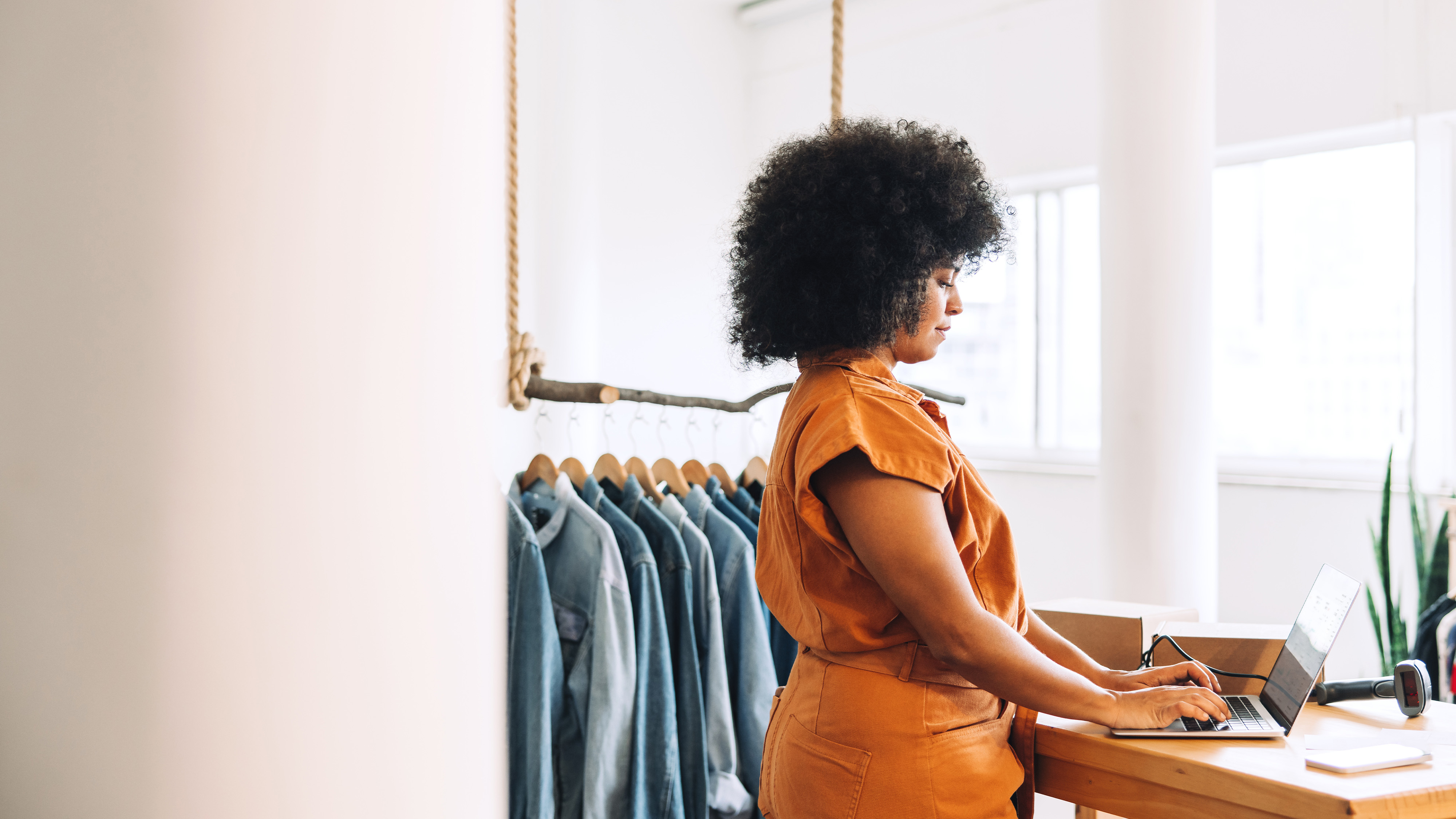 A woman working on a laptop at a clothing shop counter with a rack of denim jackets behind her